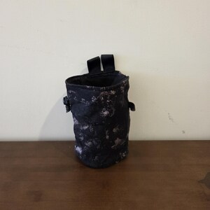 Pode incluir: Um saco de giz preto e branco salpicado para escalada. O saco tem formato cilíndrico com fecho de cordão e duas presilhas pretas na parte superior. Uma pequena presilha preta está em cada lado do saco.