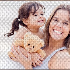 May include: A woman and a young girl are smiling and holding a brown teddy bear. The woman is wearing a light gray tank top and the girl is wearing a white tank top.
