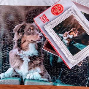 以下が含まれることがあります： 茶色と白のオーストラリア・シェパードの犬が描かれた1000ピースのジグソーパズル。パズルの箱は赤と白で、「1000 Pieces Puzzle」と「Love & Land Gifts and More」の文字が書かれています。
