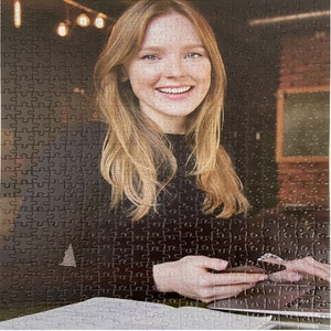 May include: A woman with long blonde hair is sitting at a table. She is wearing a black sweater and is looking at the camera. She is holding a phone in her hands.
