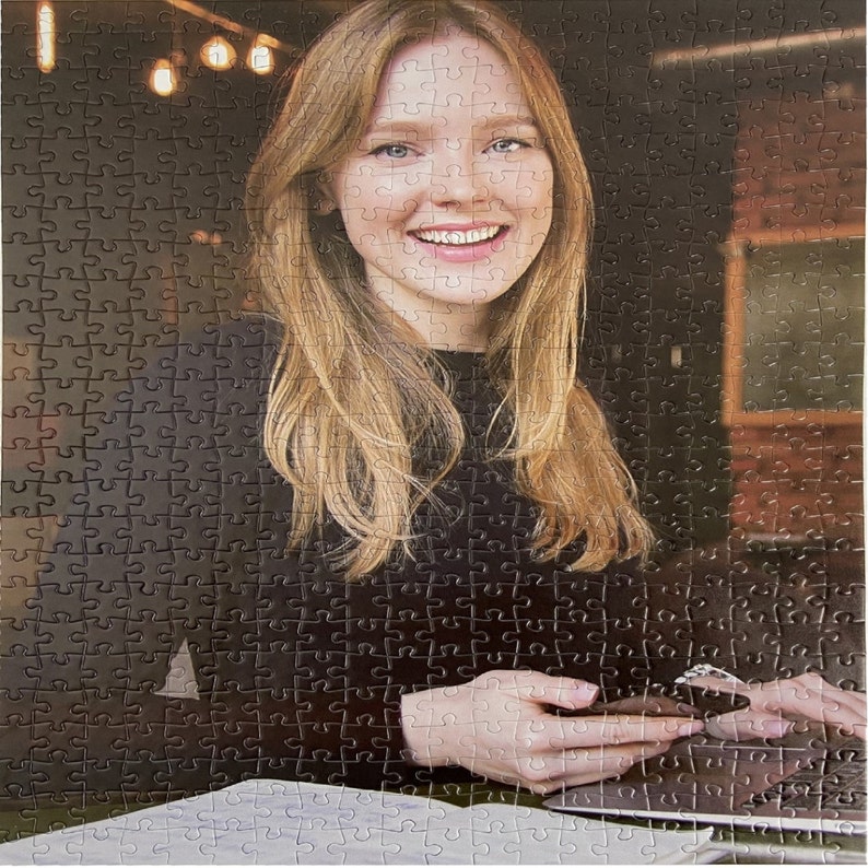 May include: A woman with long blonde hair is sitting at a table. She is wearing a black sweater and is looking at the camera. She is holding a phone in her hands.
