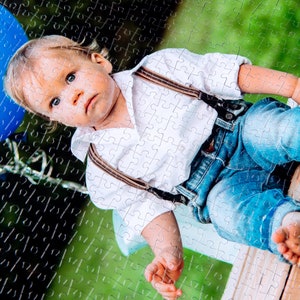 Peut inclure: Un jeune enfant portant une chemise blanche avec des bretelles marron et un jean bleu est assis sur une surface en bois. L'enfant a les cheveux blonds et regarde la cam&eacute;ra.