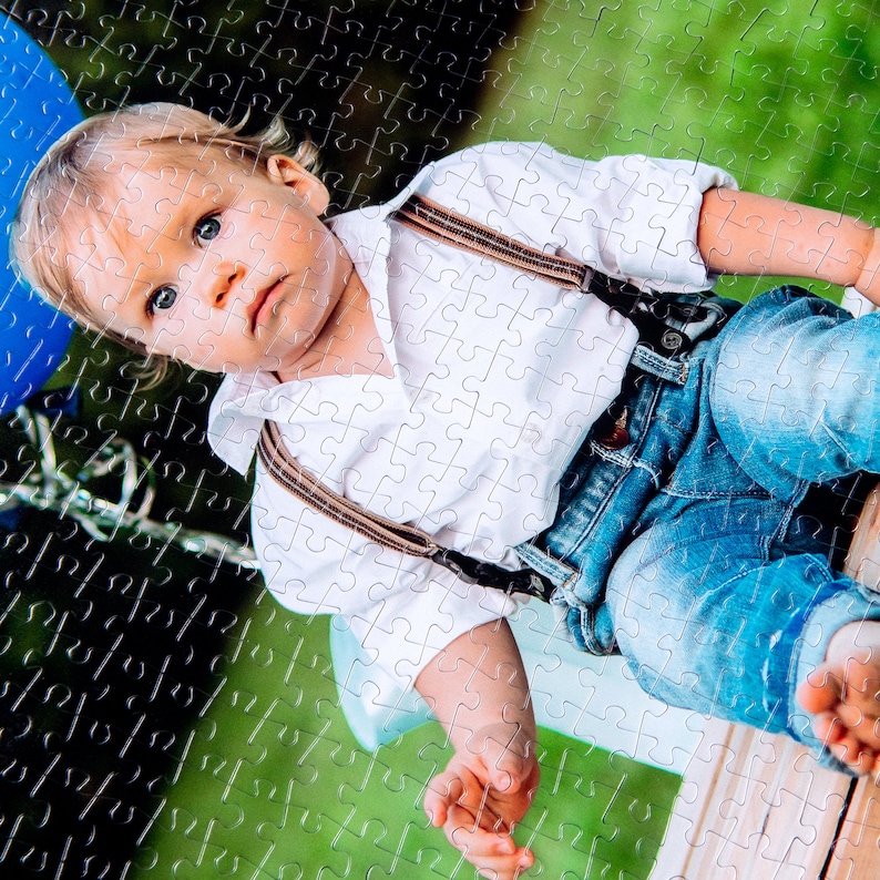 Peut inclure: Un jeune enfant portant une chemise blanche avec des bretelles marron et un jean bleu est assis sur une surface en bois. L'enfant a les cheveux blonds et regarde la cam&eacute;ra.