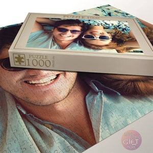 Puede incluir: Un rompecabezas de 1000 piezas con una pareja sonriendo y usando gafas de sol. La caja es beige con detalles dorados y el texto "Puzzle 1000 48x68 cm" impreso en el lateral.