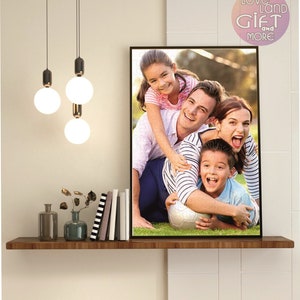 Puede incluir: Una foto enmarcada de una familia de cuatro personas sonriendo y jugando con un bal&oacute;n de f&uacute;tbol en un campo de hierba. La foto est&aacute; colgada en una pared con un estante debajo. El estante tiene un jarr&oacute;n con flores secas, una botella y una pila de libros.