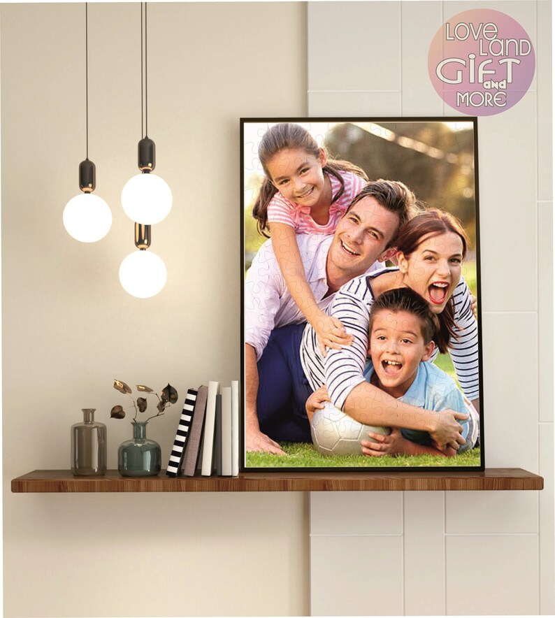 Puede incluir: Una foto enmarcada de una familia de cuatro personas sonriendo y jugando con un bal&oacute;n de f&uacute;tbol en un campo de hierba. La foto est&aacute; colgada en una pared con un estante debajo. El estante tiene un jarr&oacute;n con flores secas, una botella y una pila de libros.