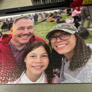 Puede incluir: Un rompecabezas familiar con un hombre, una mujer y una ni&ntilde;a. El rompecabezas est&aacute; hecho de piezas blancas con una imagen colorida de la familia impresa en la parte superior. El rompecabezas est&aacute; ensamblado sobre una superficie negra.