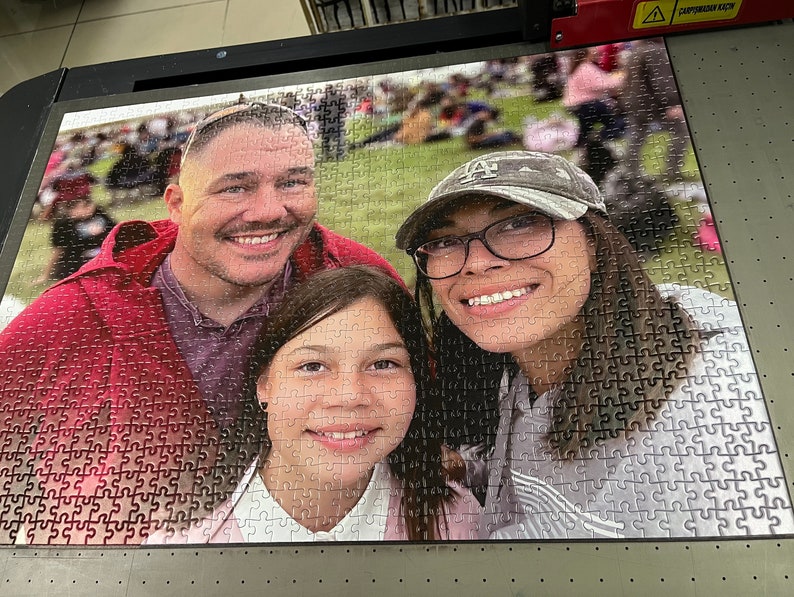 Puede incluir: Un rompecabezas familiar con un hombre, una mujer y una ni&ntilde;a. El rompecabezas est&aacute; hecho de piezas blancas con una imagen colorida de la familia impresa en la parte superior. El rompecabezas est&aacute; ensamblado sobre una superficie negra.