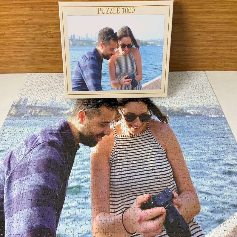 May include: A 1000 piece jigsaw puzzle featuring a couple taking a photo with a camera. The puzzle is on a white background with a blue and white water scene.