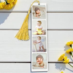 Op de afbeelding: Een witte boekenlegger met vier babyfoto's. Het heeft een gouden kwast en een klein gaatje aan de bovenkant. Gele bloemen zijn rond de boekenlegger gerangschikt.