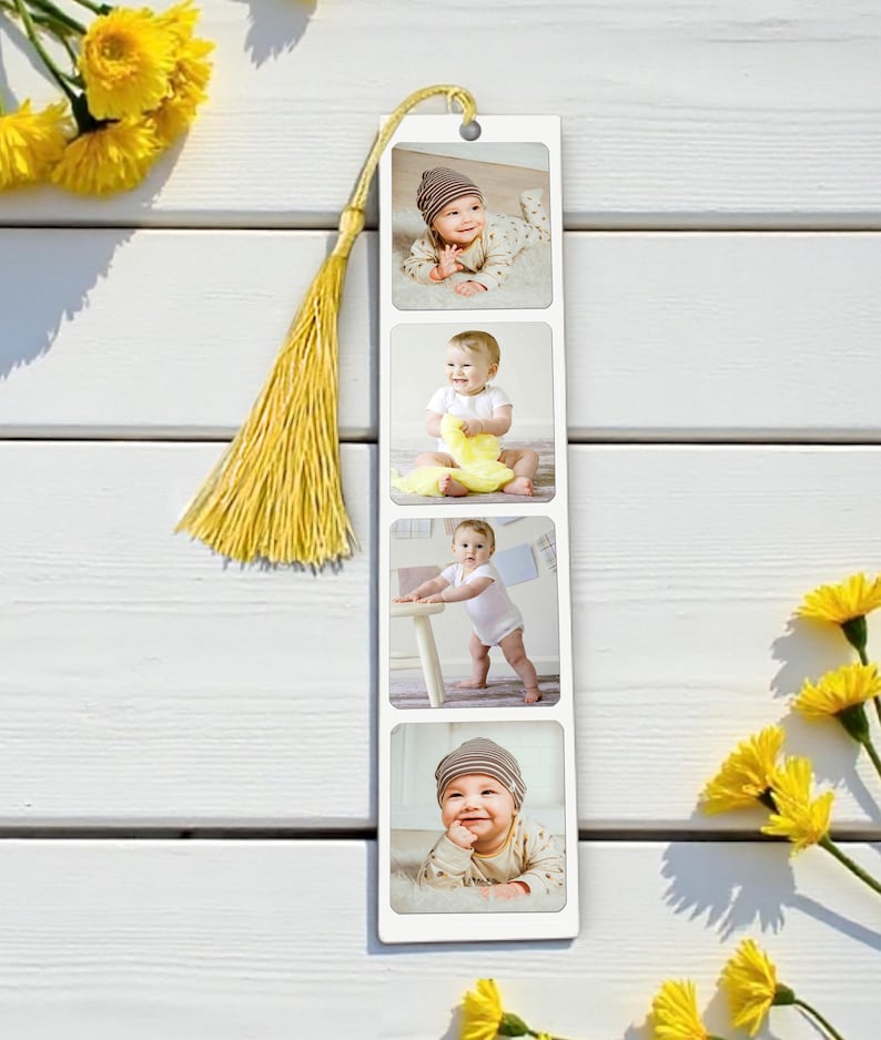 Op de afbeelding: Een witte boekenlegger met vier babyfoto's. Het heeft een gouden kwast en een klein gaatje aan de bovenkant. Gele bloemen zijn rond de boekenlegger gerangschikt.