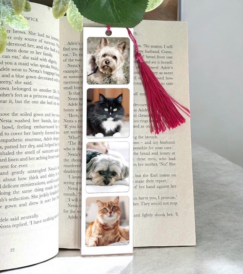 Op de afbeelding: Een rechthoekige boekenlegger met vier huisdierportretten: een Yorkshire Terrier, een zwart-witte kat, een Shih Tzu en een rode kat. De boekenlegger heeft een rode kwast en staat tussen de pagina's van een boek.