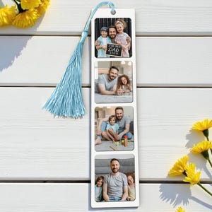 Op de afbeelding: Een witte boekenlegger met vier fotokaders, elk met een familiefoto. De boekenlegger heeft een lichtblauwe kwast en een klein gaatje aan de bovenkant. De bovenste foto bevat de tekst "THE BEST DAD EVER". Gele bloemen zijn op de achtergrond.