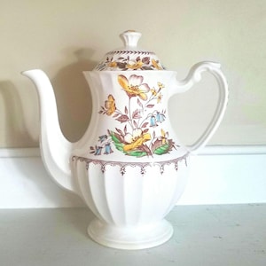 May include: White ceramic coffee pot with a floral design in shades of yellow, brown, and blue. The pot has a fluted body and a decorative border.
