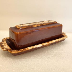 May include: A rectangular ceramic butter dish with a glossy dark brown glaze. The lid features a raised center section, while the base has integrated handles on both ends. The edges of the lid and the rim of the base are accented with a mottled cream and tan drip-style glaze pattern.