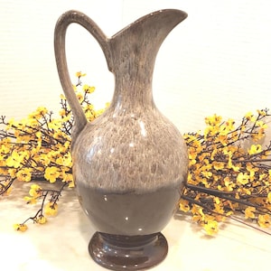 May include: A tall, two-toned ceramic pitcher with a curved handle and a flared spout. The upper portion is a speckled gray, transitioning to a solid dark brown base. The pitcher sits on a round, dark brown base. Yellow flowers are in the background.