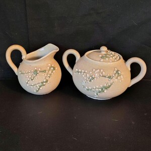 May include: A ceramic creamer and a lidded sugar bowl set in a light beige speckled finish. Both pieces feature a raised, textured floral pattern in white and sage green. The creamer has a single handle and spout, while the sugar bowl has two handles.