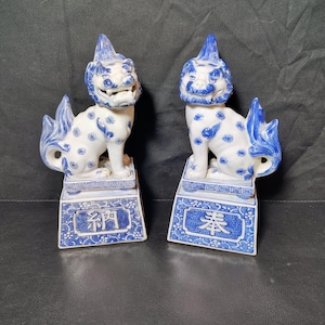 May include: A pair of white and blue ceramic Komainu lion-dog statues. Each figure features blue floral patterns on the body, a stylized mane, and sits on a rectangular base. The left base displays the kanji 納 and the right base displays 奉.