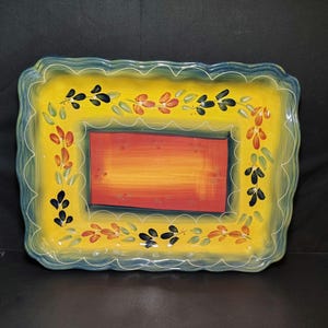 May include: A rectangular ceramic serving tray with a wavy, scalloped edge. The center features a solid orange-red rectangle bordered by a dark green line. The wide yellow rim is decorated with hand-painted leaf motifs in orange, green, and black.