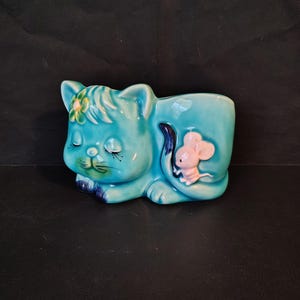 May include: A turquoise ceramic cat figurine in a sleeping position with a small pink mouse perched on its back. The cat features a painted green flower on its head, closed eyes with long eyelashes, and dark blue accents on its paws and tail.