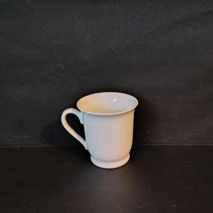 May include: A plain white ceramic mug with a smooth, glossy finish, featuring a rounded body, a flared rim, and a curved handle on the side. The mug has a small pedestal base that elevates the main body, providing a clean and simple aesthetic.