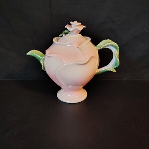 May include: A ceramic teapot shaped like a blooming rose with soft pink petals. The lid features a sculpted rosebud finial, while the handle and spout are designed to resemble green leaves. The teapot stands on a circular pedestal base.