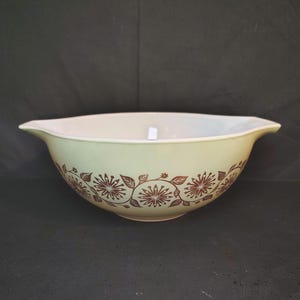 May include: A light green mixing bowl with two side handles and a brown floral pattern featuring stylized flowers and leaves encircling the exterior. The interior of the bowl is a solid off-white color, and the bowl has a smooth, rounded shape.