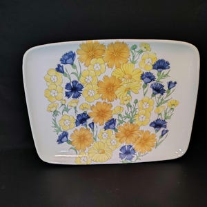 May include: A rectangular serving platter with a floral design. The flowers are yellow, orange, and blue, arranged in a circular pattern on a white background. The platter has rounded corners and a light blue border.