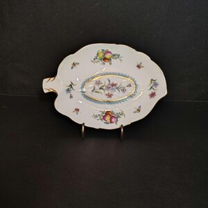 May include: White porcelain leaf-shaped dish with a gold rim and handle. The dish features a central oval design with pink and blue flowers, surrounded by fruit and butterfly motifs. A decorative piece.
