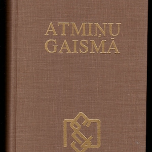 Puede incluir: Un libro marrón con el título "Atminu Gaisma" en letras doradas. Un símbolo dorado se encuentra en la parte inferior de la portada.