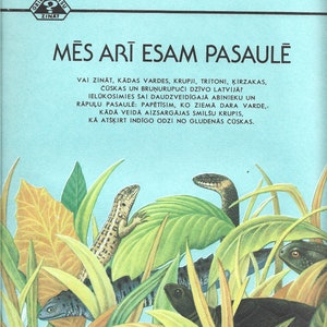 Op de afbeelding: Een boekomslag met een blauwe achtergrond en de titel "MĒS ARĪ ESAM PASAULĒ" in witte tekst. De omslag toont illustraties van een kikker, een hagedis en een slang in een groene en bruine bladerrijke omgeving.