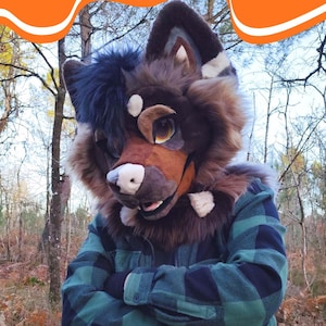 Può includere: Una testa di fursuit di lupo marrone e marrone chiaro con un accento di pelliccia nera e blu. La persona indossa una giacca a quadri verde e nera. Il fursuit ha grandi occhi espressivi.