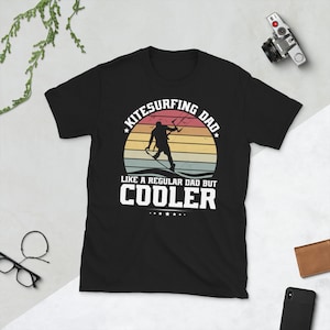 Może przedstawiać: Czarny T-shirt z białym nadrukiem przedstawiającym osobę uprawiającą kitesurfing na tle zachodu słońca. Tekst głosi "Kitesurfing Dad Like a regular dad but Cooler".