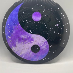 May include: A black disc with a yin yang symbol painted in purple and white. The yin yang symbol is surrounded by white dots on a black background.