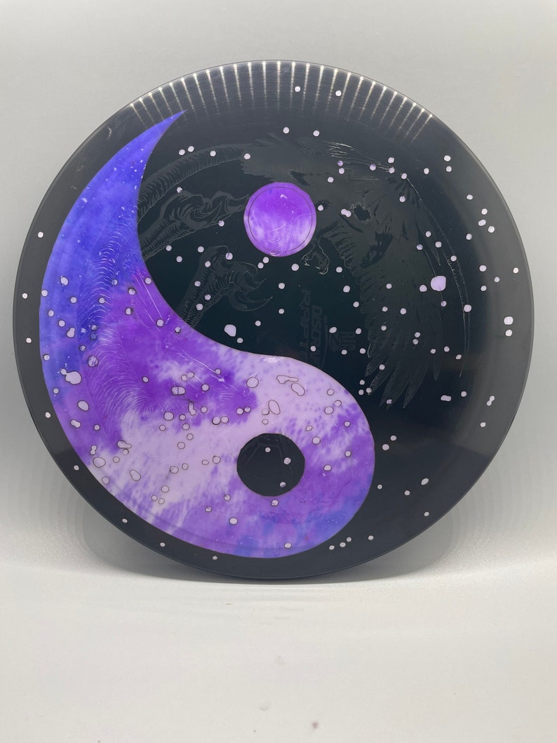May include: A black disc with a yin yang symbol painted in purple and white. The yin yang symbol is surrounded by white dots on a black background.