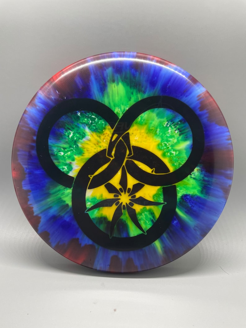 May include: A tie-dye disc golf disc with a black triquetra symbol in the center. The disc has a blue, red, green, and yellow color scheme.