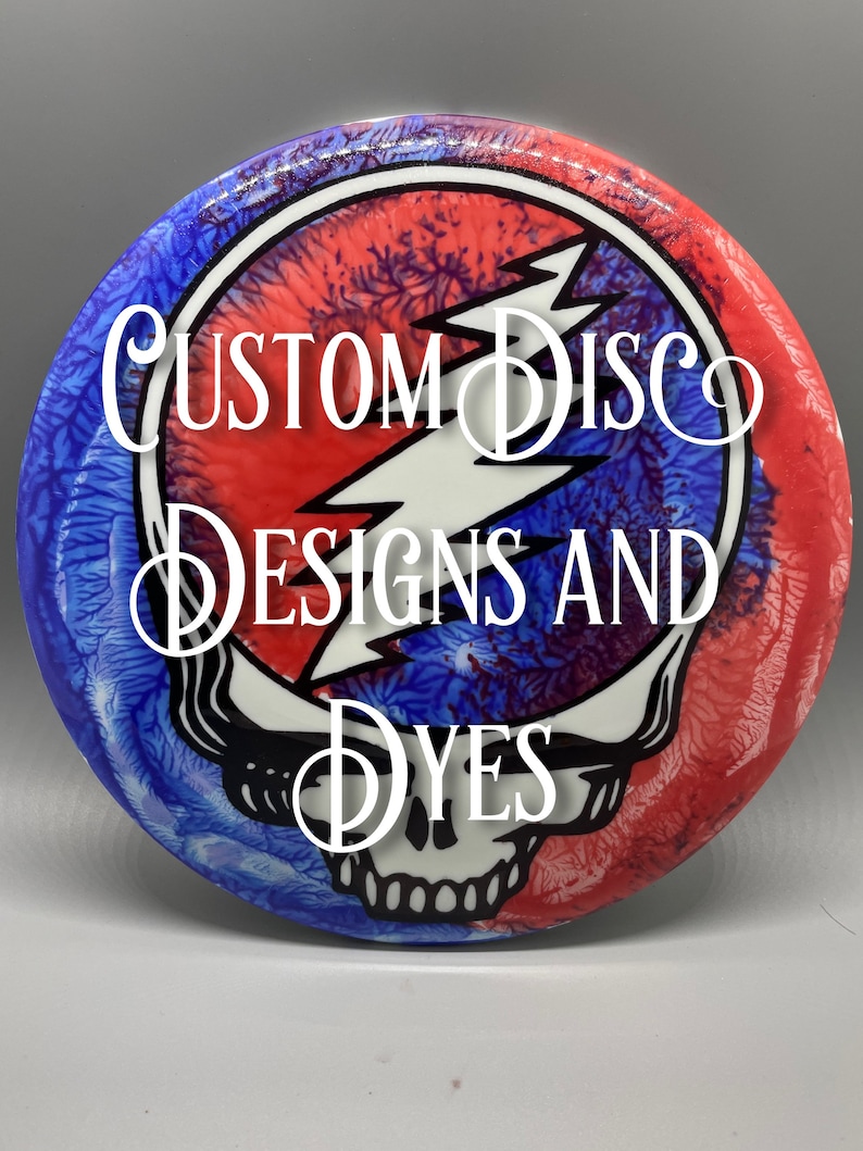 May include: A white disc with a red, white, and blue tie-dye design featuring the Grateful Dead's Steal Your Face logo. The text "Custom Disc Designs and Dyes" is written in a cursive font.