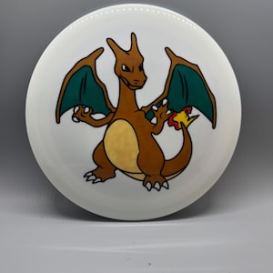 May include: A white ceramic plate featuring a cartoon Charizard design. The Charizard is depicted in shades of brown, orange, yellow, and green, with a flame at the end of its tail. The plate is round and has a glossy finish.