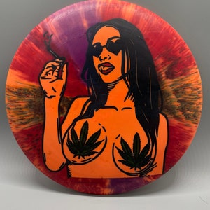 May include: A tie-dye disc with a black and white illustration of a person wearing sunglasses and holding a cigarette. The person has marijuana leaves on their chest.