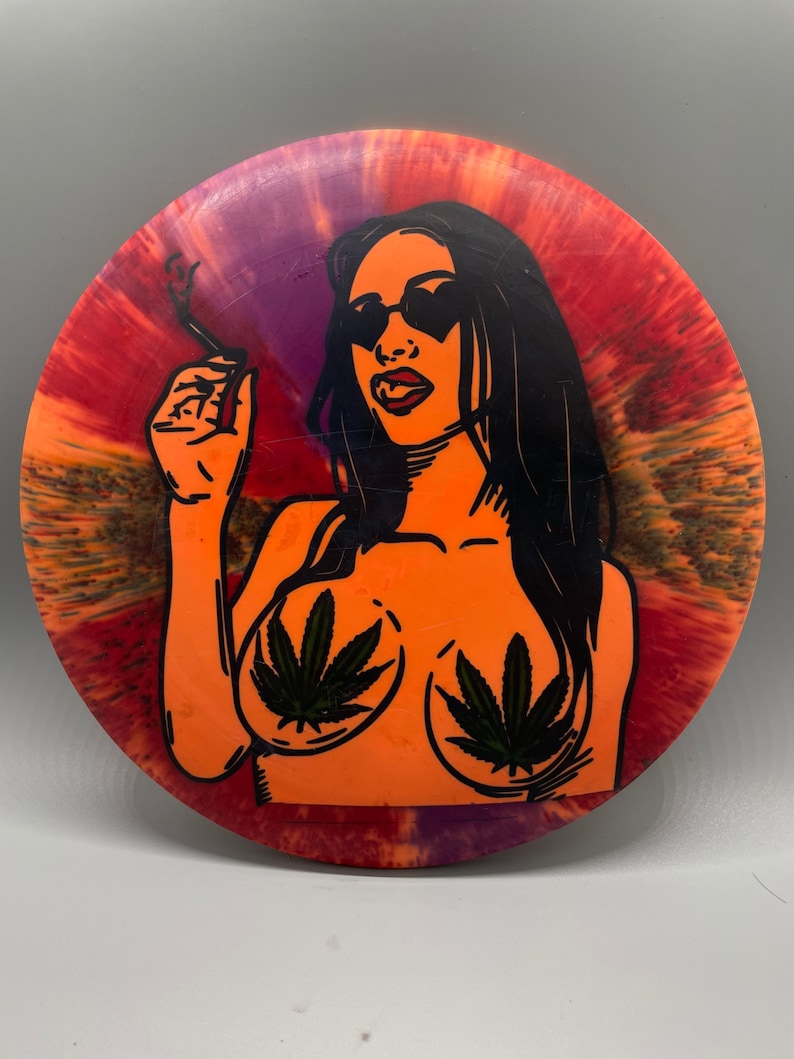 May include: A tie-dye disc with a black and white illustration of a person wearing sunglasses and holding a cigarette. The person has marijuana leaves on their chest.