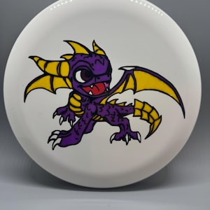 May include: A white ceramic plate featuring a cartoon dragon with purple scales, yellow wings, and a red tongue. The dragon is in a dynamic pose, with its wings spread. The plate has a smooth, glossy finish.