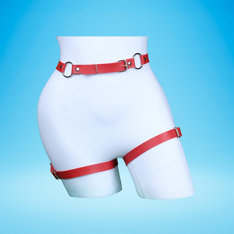 May include: A red leather body harness with silver-colored metal rings and buckles. The harness is displayed on a white mannequin against a blue background. The harness includes a waist belt and thigh straps.