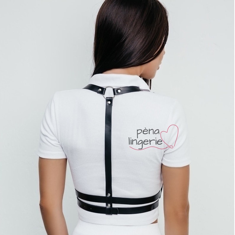 May include: A black leather harness with a silver ring and straps. The harness is worn over a white shirt with the text "p&eacute;na lingerie" and a red heart.