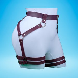 Pu&ograve; includere: Fune a forma di imbracatura in pelle bordeaux con fibbie e anelli argentati. La fune ha una cintura, cinturini per le cosce e un cinturino che passa sopra l'anca.