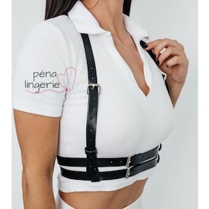 May include: A black leather harness with adjustable straps and double buckles. The harness is worn over a white shirt.