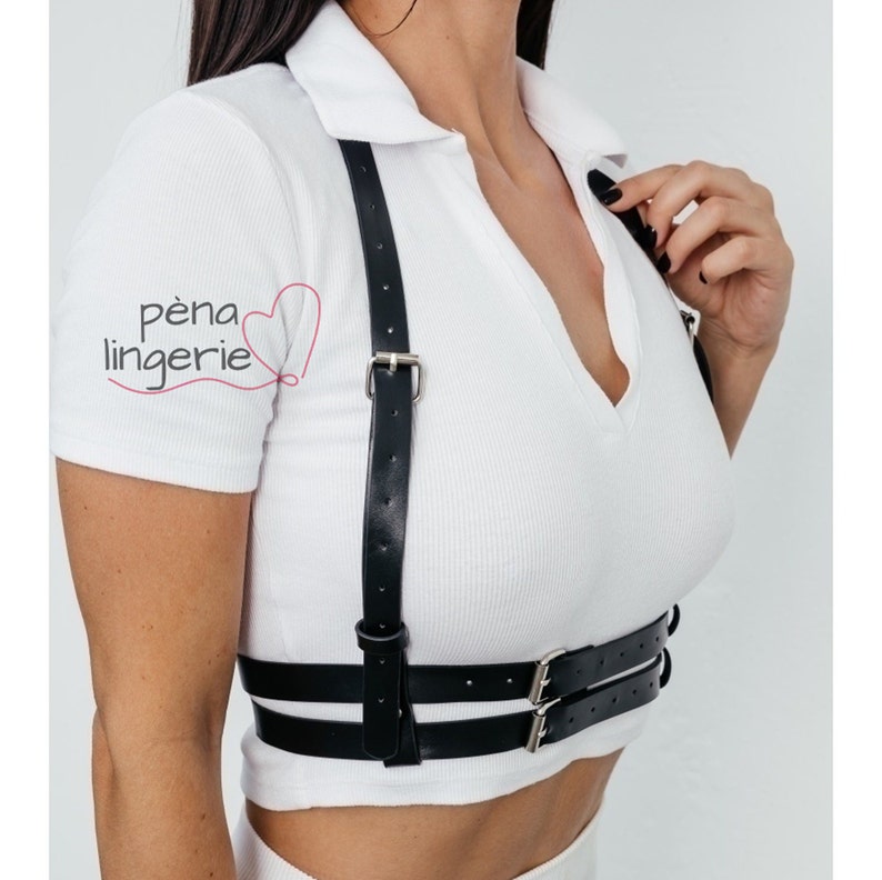 May include: A black leather harness with adjustable straps and double buckles. The harness is worn over a white shirt.