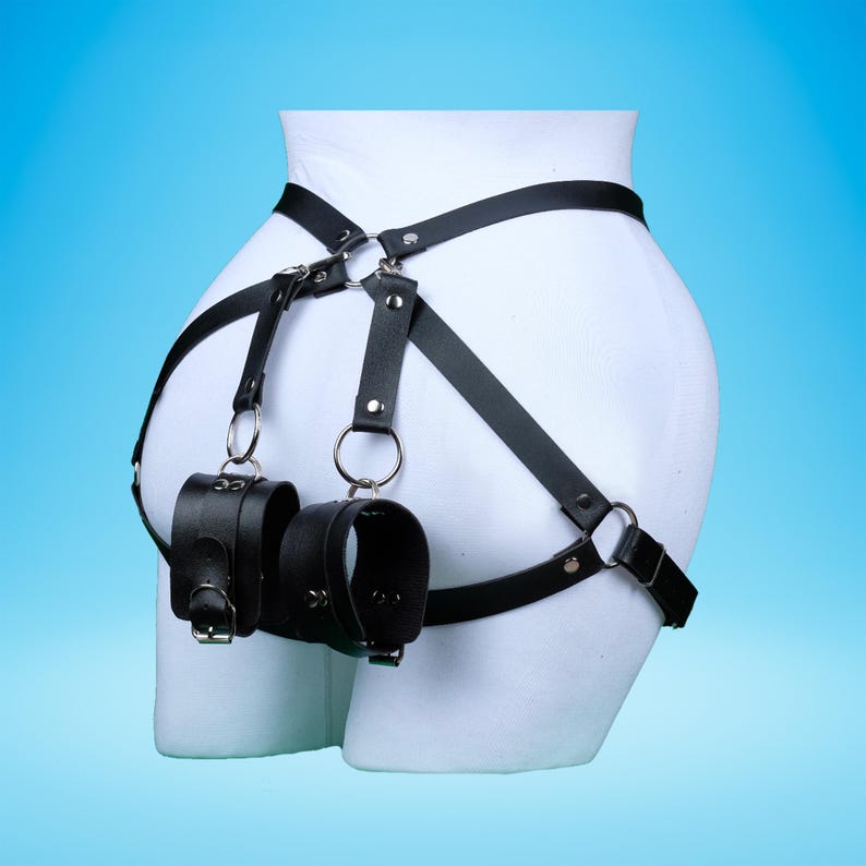 May include: Black leather body harness with attached wrist restraints. The harness features adjustable straps, metal rings, and buckle closures. The wrist cuffs are secured with buckles.