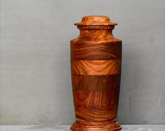 May include: A tall, wooden urn with a lid. The urn is made of various shades of brown wood, with horizontal bands of lighter and darker wood. The base and lid are rounded, and the body is cylindrical. The urn is a decorative item.