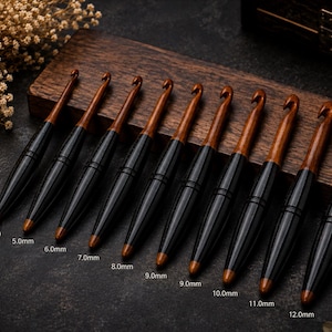 Könnte beinhalten: Ein Set aus neun Häkelnadeln mit braunen Holzschäften und schwarzen, konisch geformten Griffen. Jede Nadel ist mit ihrer Größe von 4,0 mm bis 12,0 mm beschriftet, die deutlich auf der Oberfläche aufgedruckt ist. Die Nadeln liegen auf einem Holzsockel.