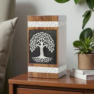 May include: A wooden urn with a tree of life design. The urn features a rectangular shape with a dark gray front panel. The tree is white and intricately cut. The top and bottom have decorative white trim. The sides are light gray.